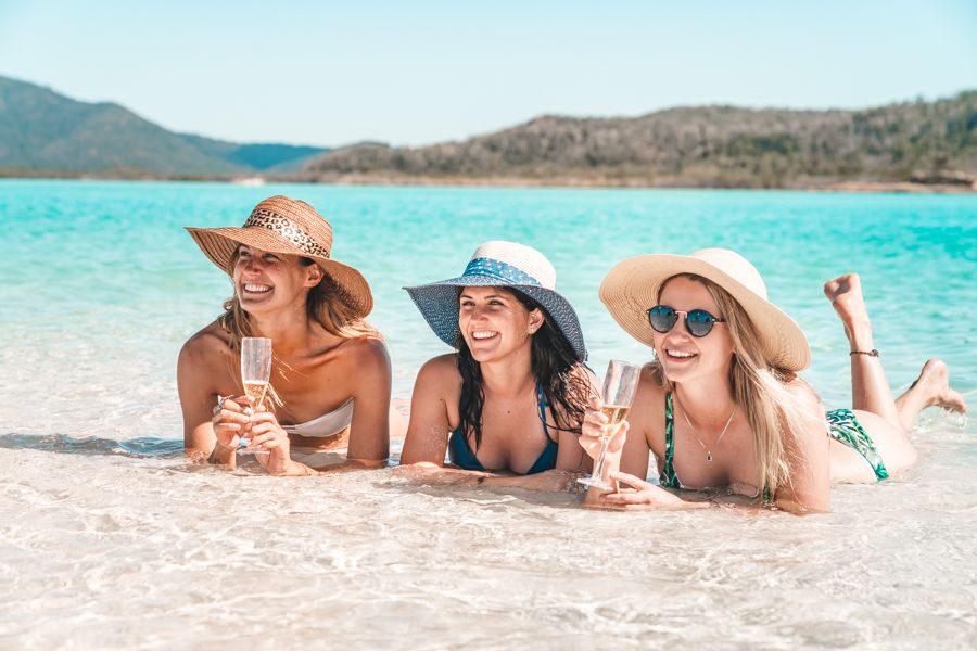 Group of three people wearing hats holding champagne, laying in the shallows at Whitehaven Beach
