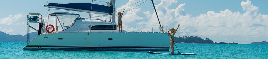 A white catamaran yacht anchored in blue water with a girl on a paddle board next to it
