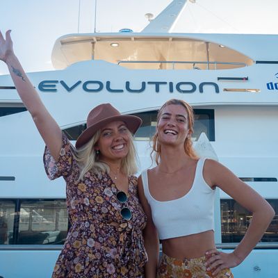 Two girls smiling in front of a boat with the sign evolutions on it 