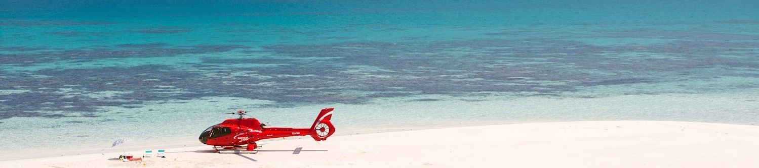 Helicopter resting on white sands of Vlasoff Cay, picnic and beach umbrella set up beside helicopter, blue waters in background