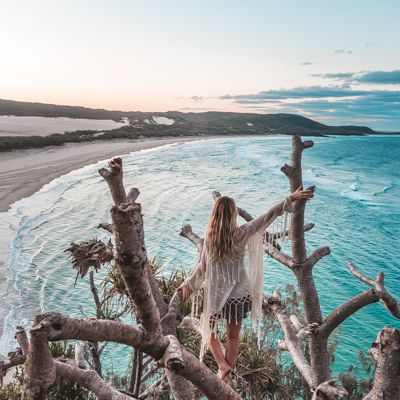 Woman in a white light coat in a tree looking out at the coastline 