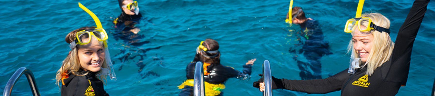 Two girls with yellow snorkels and black wetsuits on the back of a boat