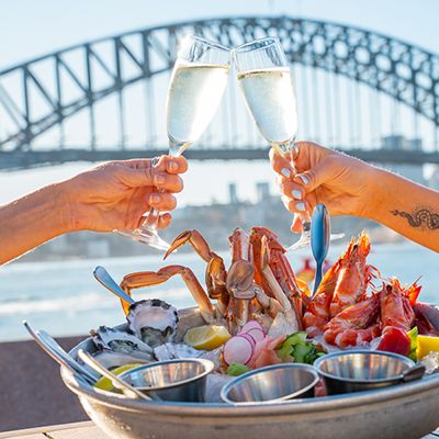 A platter of seafoods, two champagne glasses cheersing and the Sydney harbour bridge