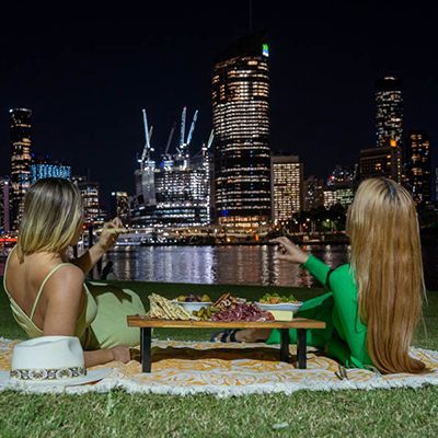 Two women on the banks of Brisbane River with a picnic at night, city in the background