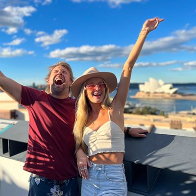 A woman with blonde hair and man in red shirt smiling in front of the Sydney Opera house