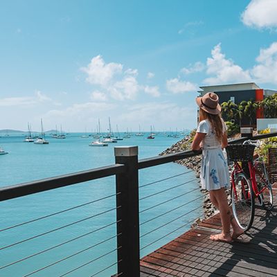 A woman in a sundress looking over the harbour with a red bike on a boardwalk
