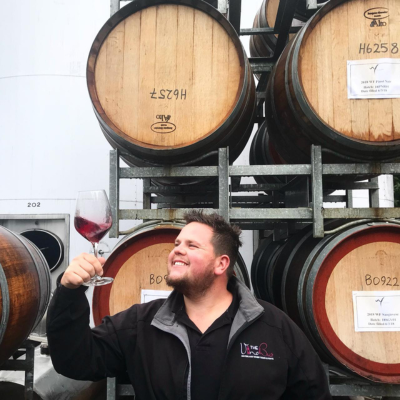 Man swirling wine in glass standing in front of wine barrels